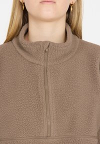 Brauner Fleece-Pullover mit hohem Kragen und halbem Reißverschluss. Der Stoff hat ein strukturiertes, weiches Aussehen und ist einfarbig ohne Muster.