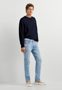 Mladý muž v námorníckom svetri, svetlomodrých džínsoch a béžových slip-on topánkach stojí s jednou rukou v vrecku na jednoduchom svetlom pozadí.