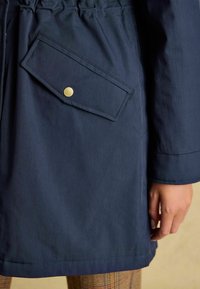 Námornícky modrá bunda so zúženým pásom, s diagonálnou chlopňovou kapsou zaistenou zlatým gombíkom a hladkou, matnou textúrou.