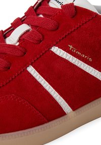 Gros plan d'une basket en daim rouge avec des bandes blanches, des lacets rouges assortis et le nom de la marque "Tamaris" sur le côté au-dessus d'une semelle beige.