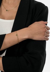 Gold bracelet with heart charm, linked design, worn on wrist against a black fabric background. Simple and sleek appearance.