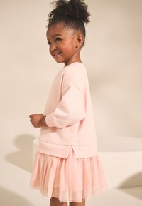 Rózsaszín, hosszú ujjú pulóver texturált, hálós rétegezett szoknyával. Puha anyag, kényelmes szabás és lekerekített szegély hátul.