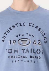 Modré tričko s krátkým rukávem vyrobené z měkké tkaniny. Obsahuje černý potištěný text a logo v různých velikostech, s pruhovaným vzorem na písmenkách.
