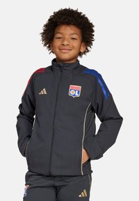 Zwarte sportjack met blauwe en rode gestreepte schouders, gouden accenten, ritssluiting en het logo van Olympique Lyonnais. Gladde textuur.
