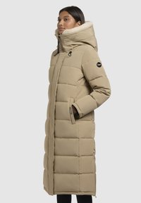 Bežs pūkains mētelis ar kapuci, ar kviltota dizaina, sānu kabatām un mīkstu, teksturētu iekšējo oderi.