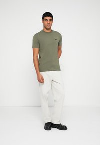 Short-sleeve olive green t-shirt with a subtle brand logo, paired with light-colored trousers and black shoes, against a plain background.