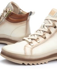 Cream leather high-top sneakers with a brown leather accent, featuring gold hardware, textured laces, and a rugged rubber sole.