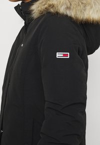 Schwarze isolierte Jacke mit einer Kapuze aus Kunstfell. Mit einem Logo-Patch in Rot, Weiß und Blau am Ärmel. Glatte Textur mit einer taillierten Form.