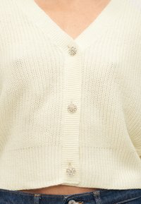 Cardigan en maille crème avec trois boutons décoratifs ornés de bijoux, porté sur un jean en denim bleu, raccourci à la taille.