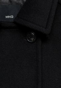 Abrigo de lana negro con una superficie texturizada, que presenta un collar clásico y un cierre de botón negro brillante. Logotipo de MNG en la etiqueta interior.