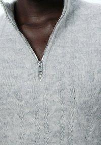 Grå kabelstickad tröja med hög krage, som har en frontdragkedja i silvermetall. Mjuk textur och detaljerad mönster synlig.