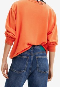 Oranje oversized sweatshirt met opgerolde mouwen, gecombineerd met donkerblauwe jeans met achterzakken en een groene stoffen label aan de tailleband.