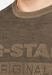 Närbild av en person som bär en brun texturerad tröja med upphöjd text "G-STAR ORIGINAL" på framsidan.