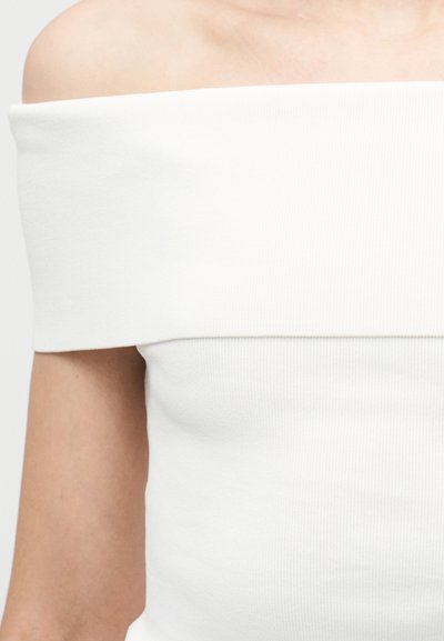 Haut blanc à épaules dénudées en maille côtelée porté par une personne, mettant l'accent sur le décolleté et la partie supérieure du torse.