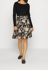 Femme portant un haut noir à manches longues, une jupe noire de longueur genou avec un motif floral beige et rose, et des chaussures noires à enfiler, se tenant sur un fond blanc.