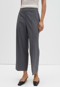 Grijze wijde broek met hoge taille, voorplooien en strakke lijnen, gecombineerd met zwarte pumps met puntige neus op een neutrale achtergrond.