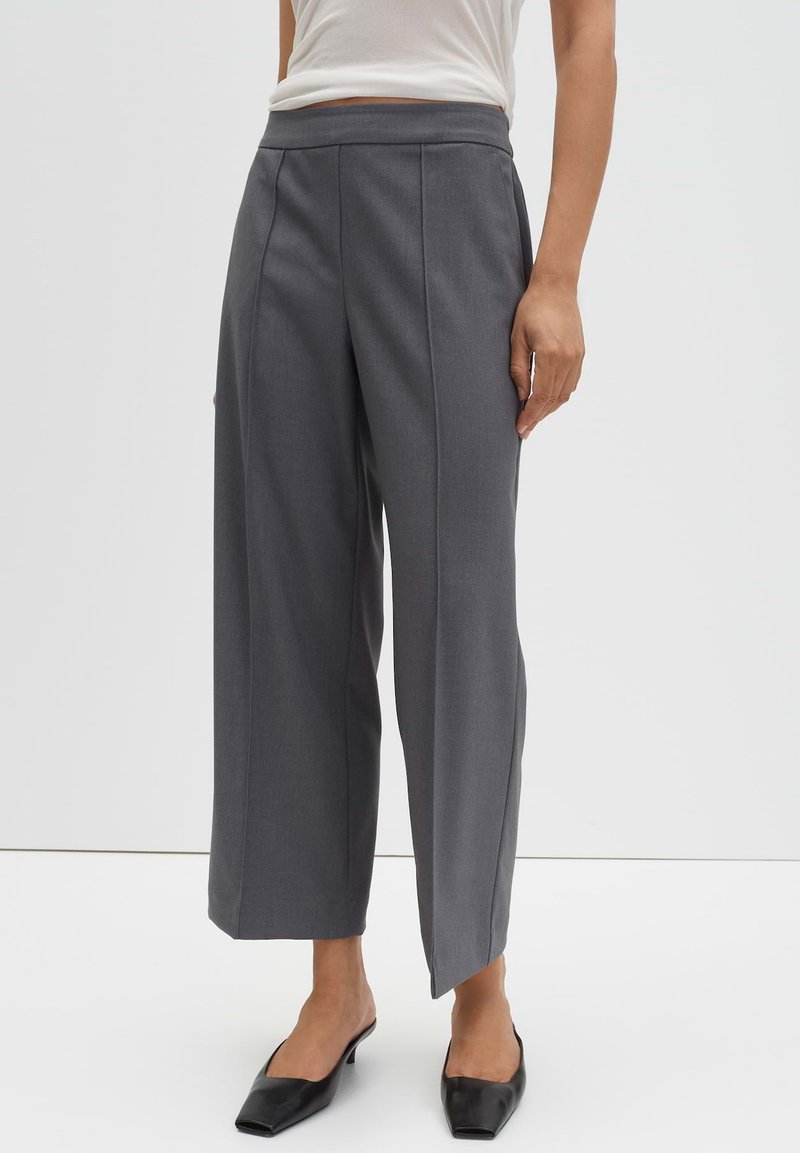 Grijze wijde broek met hoge taille, voorplooien en strakke lijnen, gecombineerd met zwarte pumps met puntige neus op een neutrale achtergrond.