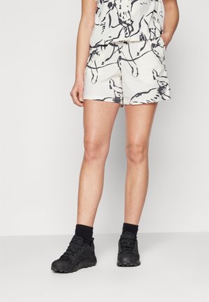 Vrouw draagt witte shorts met zwarte abstracte print, gecombineerd met zwarte sokken en zwarte sportschoenen, staand voor een egale achtergrond.