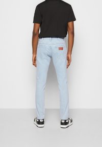 Vaqueros de color azul claro con corte ajustado, que cuentan con bolsillos traseros y una parche de cuero marrón sobre el bolsillo derecho. Llevados con una camisa negra y zapatillas.