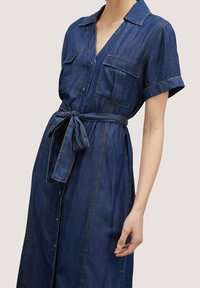 Denimskjortklänning i mörkblått med korta ärmar, två bröstfickor, en V-ringning och ett midjeband för en figurnära look. Slen yta.