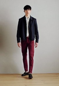 Schwarze Bomberjacke über einem weißen Hemd, kombiniert mit maroonfarbenen Slim-Fit-Hosen und schwarzen Schuhen, vor einem neutralen Hintergrund.