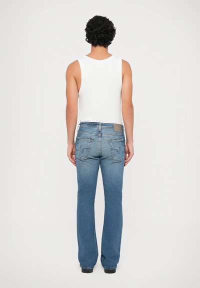 Blaue Jeans mit ausgestelltem Bein und mittelhoher Taille, ausgestattet mit zwei Gesäßtaschen und einem hellbraunen Lederpatch am Bund. Dazu wird ein weißes Tanktop getragen.