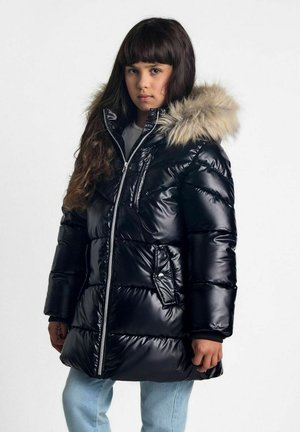 Schwarze Pufferjacke mit glänzender Optik, mit einem mit Pelz gefütterten Kapuzen und Reißverschluss, sowie mit Reißverschlusstaschen, getragen über hellblauen Jeans.