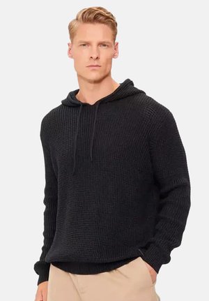 Blonde man met een zwarte gebreide capuchontrui en beige broek, staat met één hand in zijn zak en kijkt vooruit.