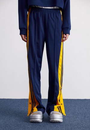 Persona que lleva pantalones deportivos azul marino con rayas laterales amarillas y zapatillas plateadas, de pie sobre una alfombra azul.