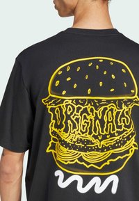 T-shirt nera con un grande graphic di un hamburger giallo sul retro, con la parola "ORIGINALS" in un testo stilizzato e accenti bianchi ondulati sotto.