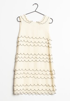 Robe crème sans manches avec bordure noire festonnée au col et rangées horizontales, suspendue à un cintre noir sur fond blanc.