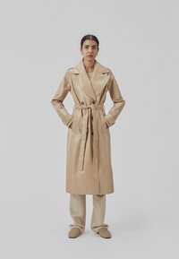 Beige, dobbeltbrystet trench coat med bælte i taljen, lavet af skinnende patenteret materiale. Har brede klapper og lange ærmer.