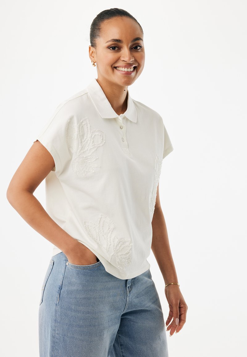 Biała koszulka polo z krótkim rękawem, zdobiona teksturowanym kwiatowym haftem. Połączona z jasnoniebieskimi jeansami o luźnym kroju.