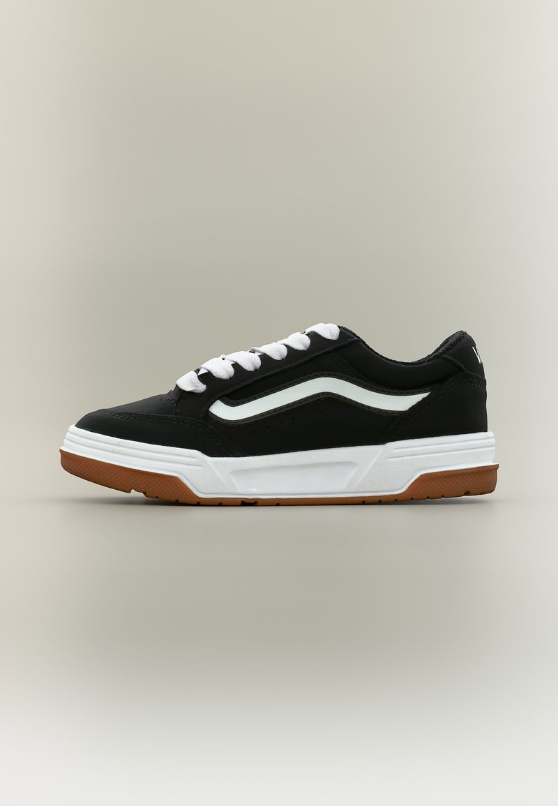 Μαύρο αθλητικό παπούτσι χαμηλού προφίλ με λευκά κορδόνια, λευκή πλευρική ρίγα, παχύ λευκό πέλμα και καφέ πέλμα, εμφανισμένο σε πλευρική όψη σε ουδέτερο φόντο.