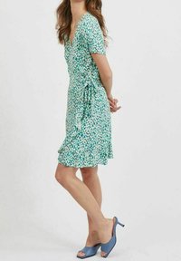 Blommig omlottklänning i grönt och blått med korta ärmar, V-ringning, justerbart knytband i midjan och en utsvängd nederdel. Matchad med blåa slip-on skor.