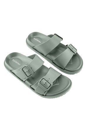 Een paar grijze instapsandalen met twee verstelbare gespen en gestructureerde zolen, met het merk "holster" op de voetbedden.