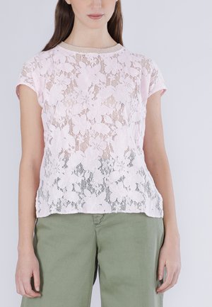 Donna che indossa una maglia di pizzo floreale rosa chiaro a maniche corte e pantaloni verde oliva, in piedi davanti a uno sfondo semplice.