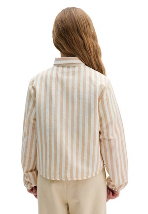 Person mit langen Haaren steht mit dem Rücken zum Betrachter, trägt ein beiges und weißes, vertikal gestreiftes Langarmhemd und beige Hose.