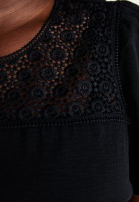 Black fabric top featuring a scalloped lace yoke with floral embroidery, showcasing intricate detailing and a smooth texture.