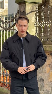 Giacca imbottita nera con ampio colletto e tasche anteriori, dotata di chiusura con zip e una piccola tasca laterale con zip, indossata sopra una camicia bianca.