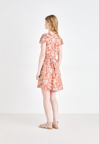 Blumenkleid mit kurzen Ärmeln, das einen orangefarbenen Hintergrund und weiße Blätter hat. Taillengürtel, ausgestellter Rock und flache Sandalen.