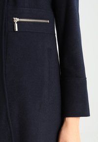 Navy wool coat with a zippered chest pocket, soft texture, and structured sleeves, featuring a tailored fit and minimalistic design.