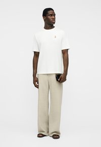 Άνδρας που φοράει λευκό πουκάμισο με κοντά μανίκια και μικρό λογότυπο, μπεζ παντελόνι με φαρδιά μπατζάκια, μαύρα σανδάλια, κρατώντας ένα μαύρο τηλέφωνο, στέκεται μπροστά σε ένα απλό φόντο.