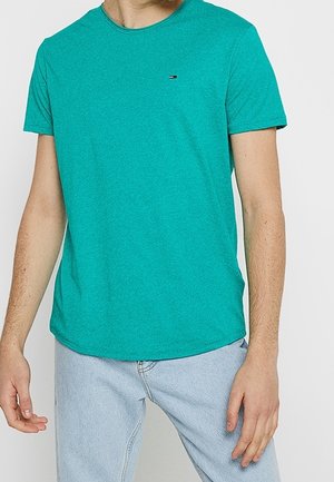 T-shirt vert canard à manches courtes en tissu doux, avec un col rond et un petit logo sur la poitrine. Porté avec un jean bleu clair.