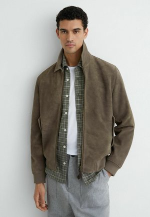 Mladý muž oblečený v hnedej semišovej bunde cez zelenú károvanú košeľu a biele tričko, stojaci pred jednoduchou svetlou stenou.