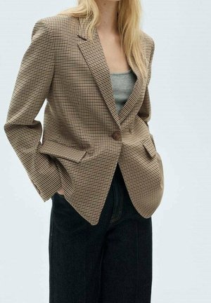Woman wearing brown houndstooth blazer with button, grey tank top, and black pants, hands in pockets, against plain light background.