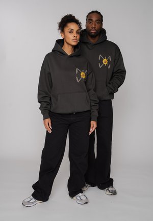 Zwei Personen tragen schwarze Kapuzenpullover mit Sonnenblumen-Logos, stehen dicht beieinander und blicken nach vorne vor einem einfarbigen grauen Hintergrund.