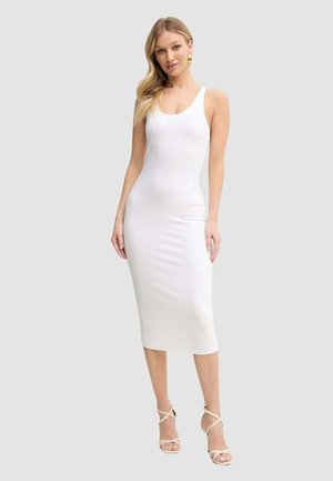 Donna che indossa un vestito midi bianco aderente senza maniche e sandali beige con tacco alto e cinturini, in piedi davanti a uno sfondo chiaro uniforme.