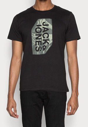 Mężczyzna nosi czarną koszulkę z krótkim rękawem z metaliczną, srebrną naszywką i dużym, czarnym napisem "JACK & JONES" z przodu.