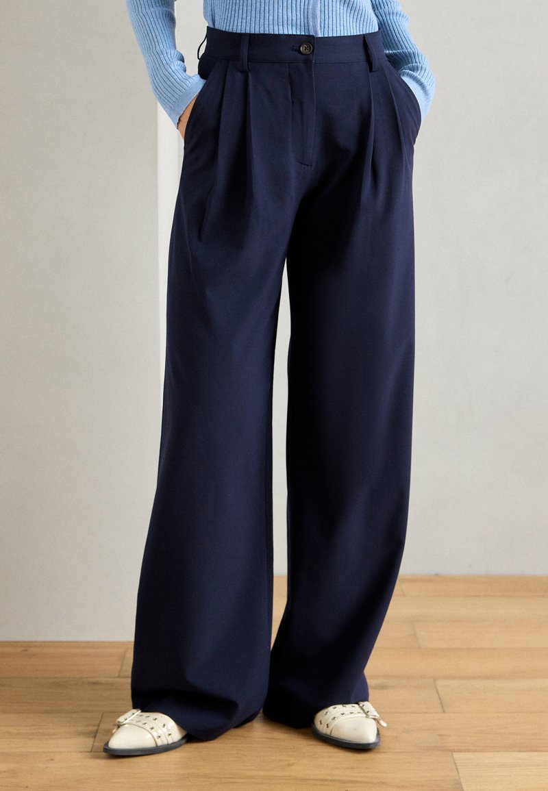 Person, der eine marineblaue weite Hose, einen hellblauen gerippten Pullover und weiße mit Nieten verzierte Schuhe trägt, steht auf einem Holzboden.
